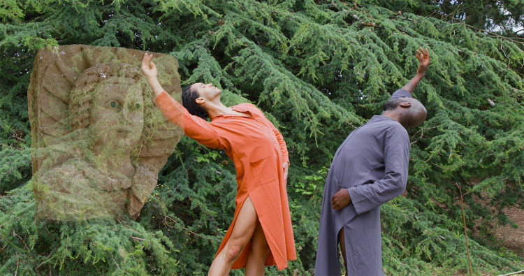 An outdoor scene with two dancers stretching backwards in unison near the cedar tree. The stone sculpture remains superimposed on the left side.