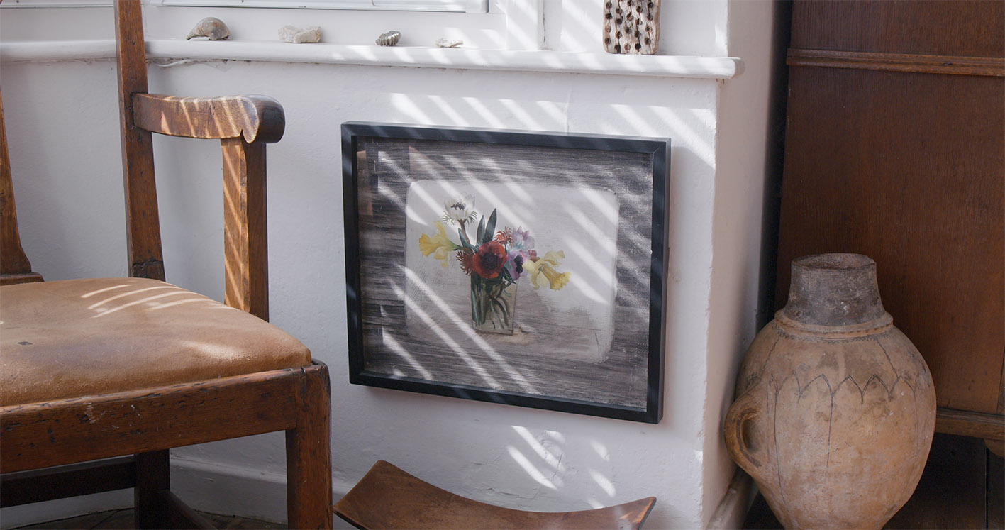 A framed painting of a flower arrangement featuring red, yellow, and white blooms is leaning against a white wall near a wooden chair and a clay pot.