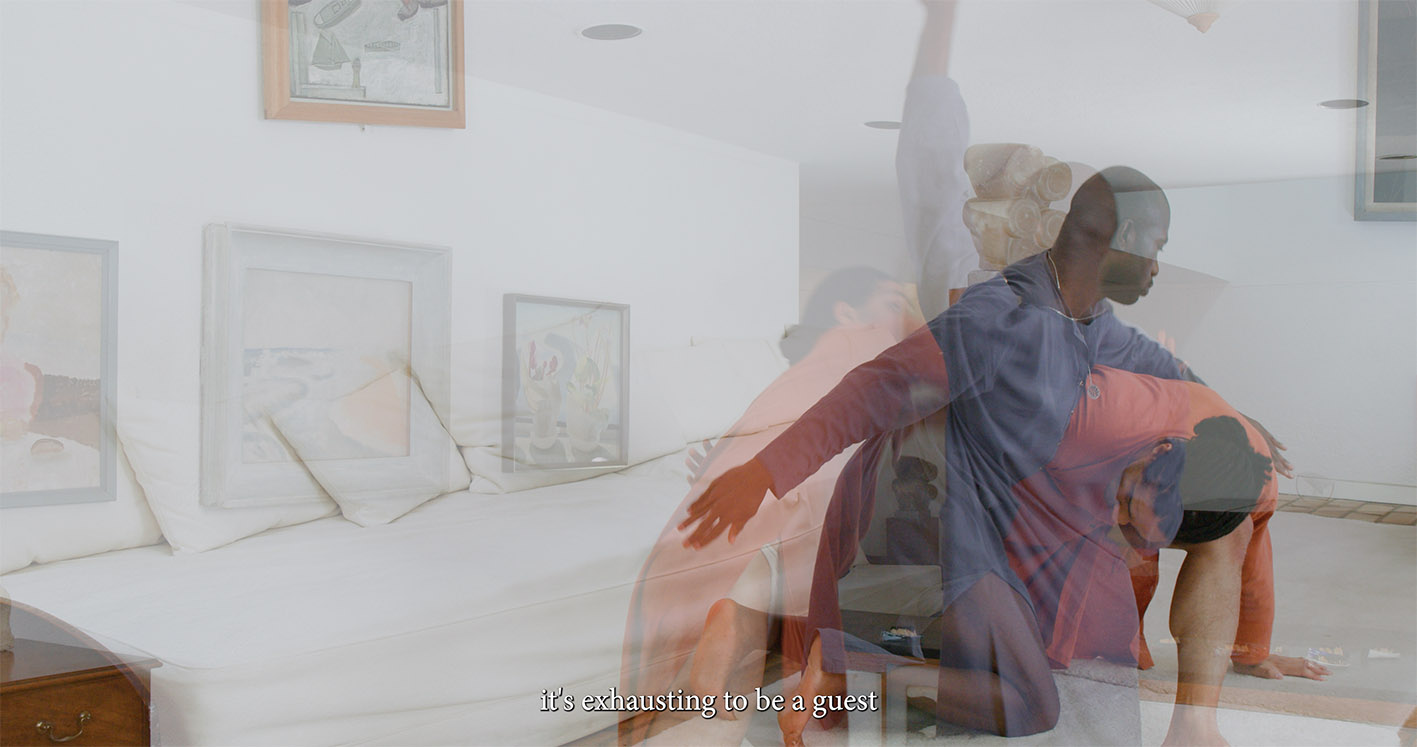 Two dancers, one in blue and one in orange, are captured mid-performance in a room with a white sofa, framed artwork, and a large clay sculpture in the background. Text at the bottom reads: “it’s exhausting to be a guest.”