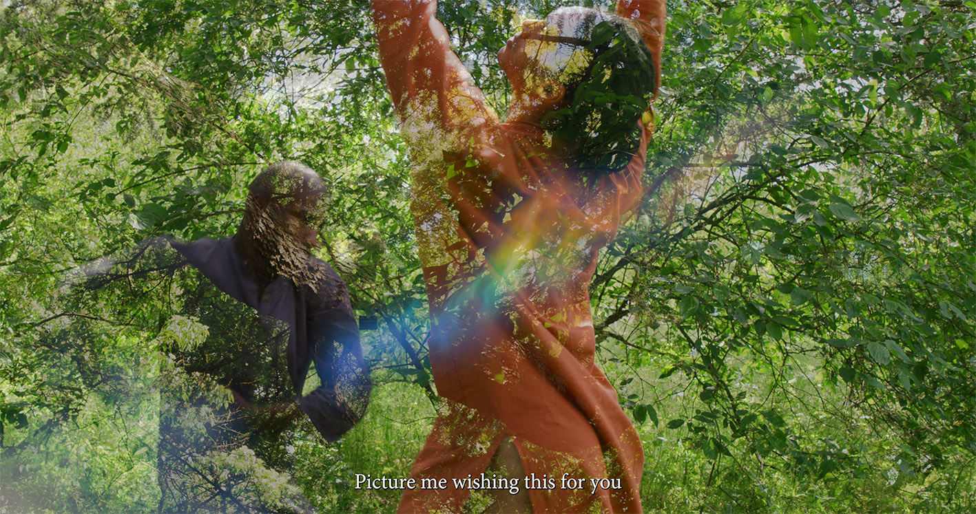 Two dancers in bright outdoor greenery, one wearing orange and the other blue, are captured mid-movement with their arms raised. Sunlight filters through the leaves. Text at the bottom reads: “Picture me wishing this for you.”