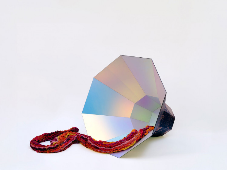 A large, iridescent megaphone-shaped object with a thick, multi-coloured plaited fabric spilling from the inside in shades of red, pink, orange, and yellow with beaded chain.