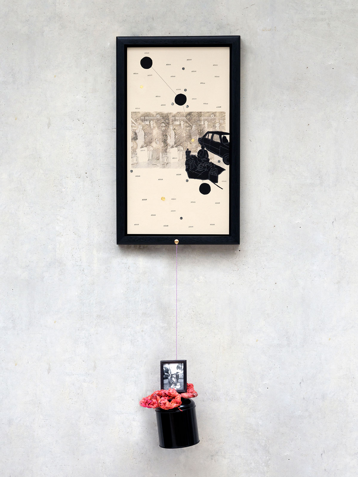 A framed artwork hangs on a gallery wall, featuring a collage of translucent drawings, silhouetted figures, circular shapes, lines, and the recurring word 'Asian.' It is paired with a hanging metal sculpture that includes a red plaited fabric flower and a miniature frame.