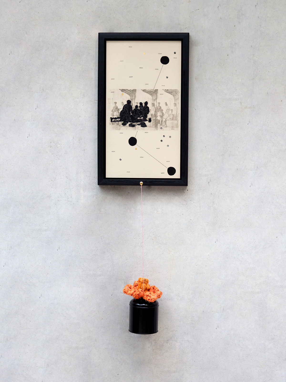 A framed artwork hangs on a gallery wall, featuring a collage of translucent drawings, silhouetted figures, circular shapes, lines, and the recurring word 'Asian.' It is paired with a hanging metal sculpture that includes an orange plaited flower and a miniature teddy bear.