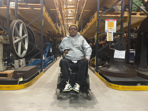 Christopher Samuel sat in the Birmingham Museum Trust Museum Collection Centre. Christopher is sat in a wheelchair wearing a grey hoodie and black trousers. Behind him, huge racks of artefacts stretch off into the distance.