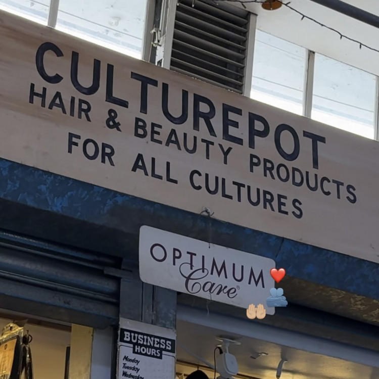 A photo of a sign for a hair and beauty shop. The sign reads ‘Culturepot Hair & Beauty Products For All Cultures’. Underneath this sign there is a smaller one which says ‘Optimum Care’. On top of the photo, around the ‘Optimum Care’ sign there is a red love heart emoji, an emoji of people hugging and an emoji of two open hands, palms upwards.