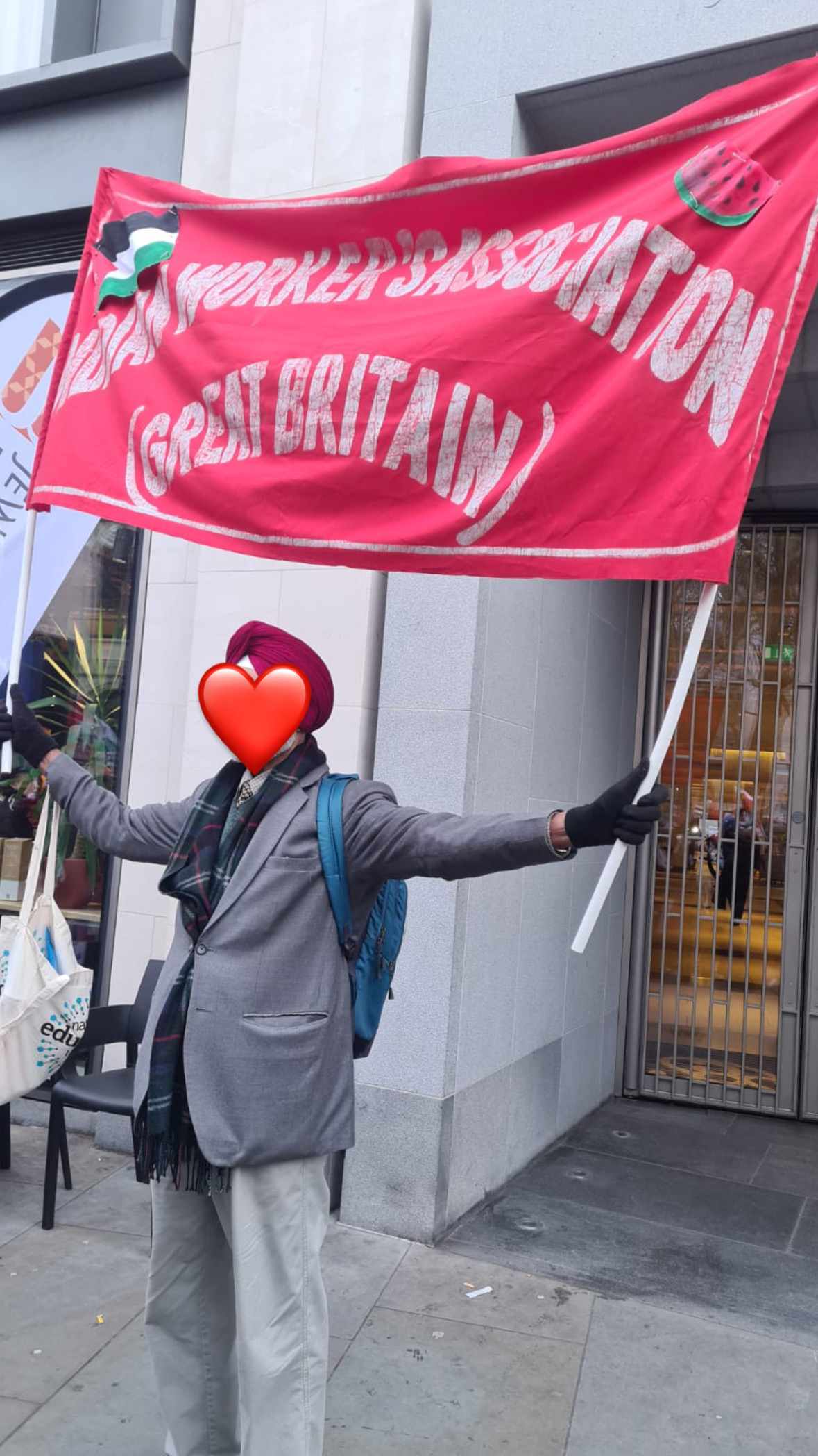A man at a march for Palesinte. He is holding a big red banner. There is white text on the banner which reads ‘Indian Workers Association (Great Britain)’. Also on the banner is a Palestinian flag and a watermelon.