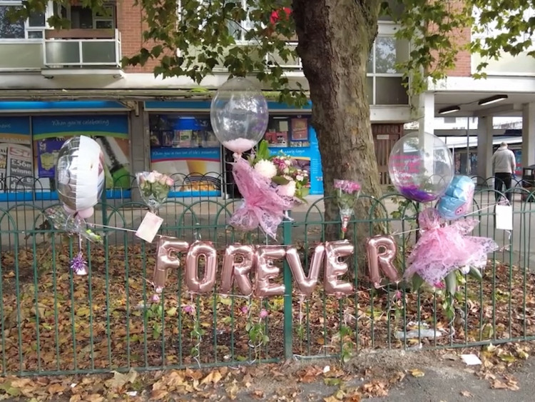 A picture of a public memorial in front of some shops and a tree. The memorial consists of pink balloons that spell out the word ‘forever’, more balloons and bouquets of flowers.