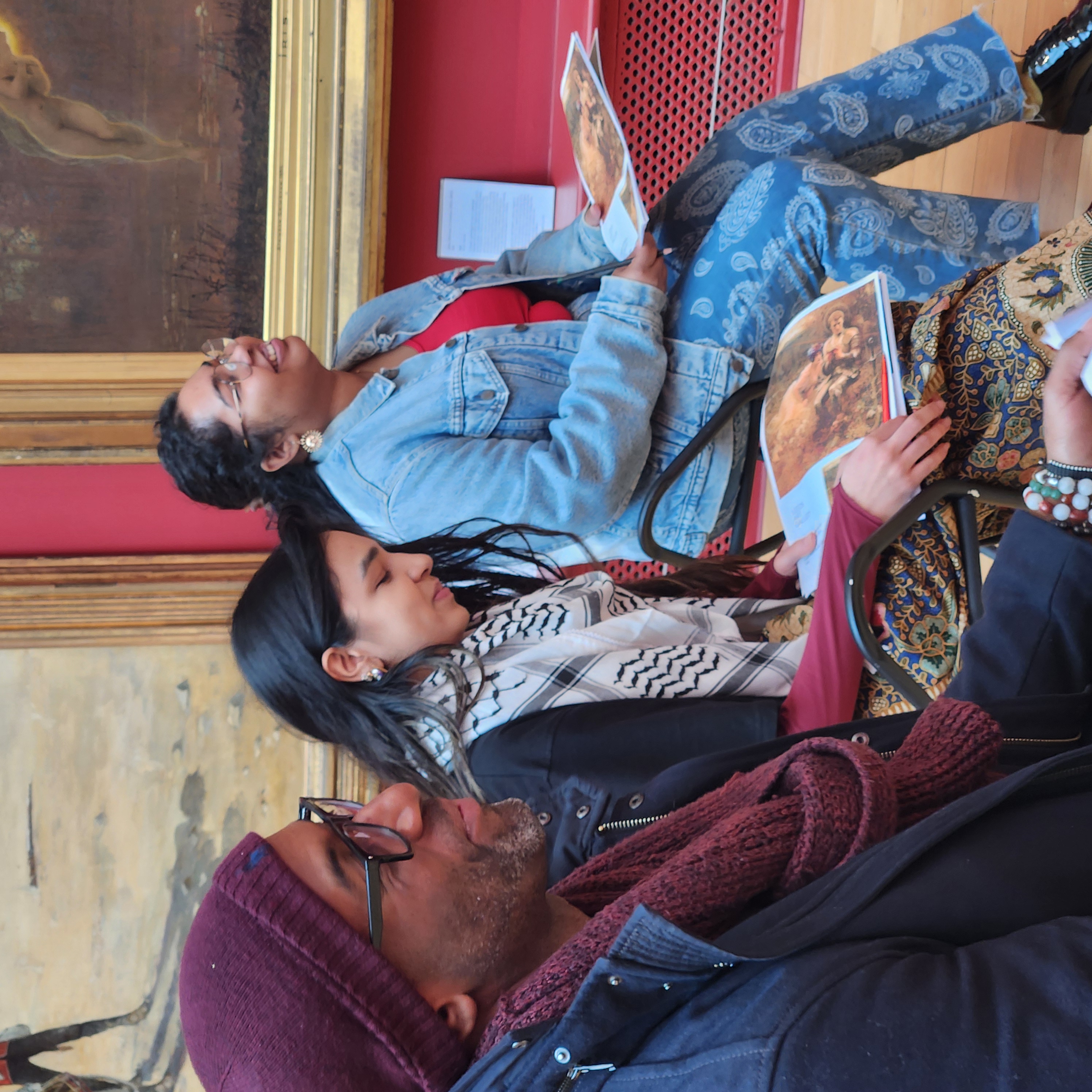 A group of people sit in a gallery room with red walls and framed paintings. Some participants hold sketchbooks and pencils, engaging in a drawing activity while others smile and interact.