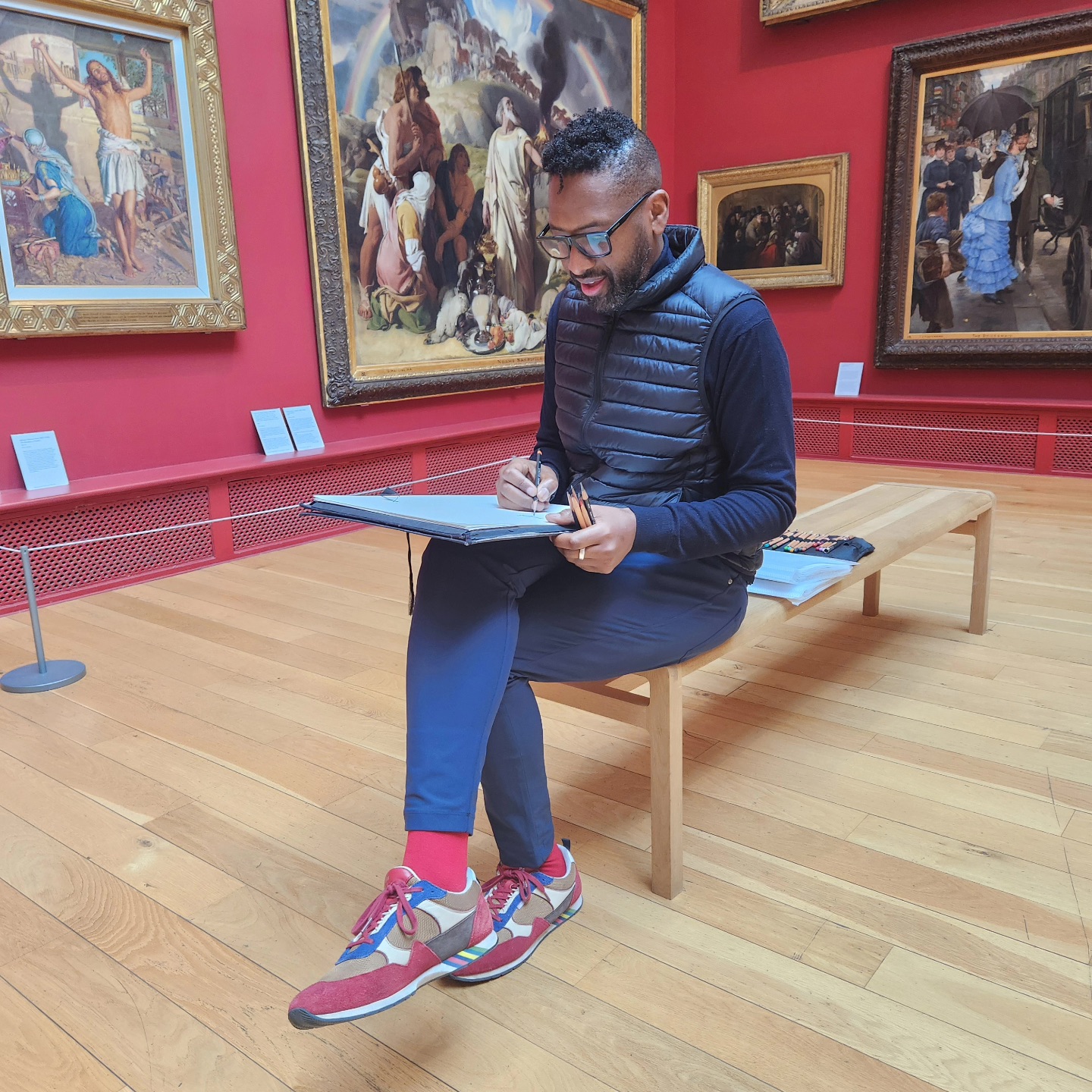 An artist sits on a wooden bench in a gallery room with red walls, surrounded by large framed paintings. The artist, wearing glasses, a black vest, and red and beige trainers, holds pencils and actively works on a sketchpad balanced on their lap. The scene is calm and focused, with the classic artwork in the background complementing the setting