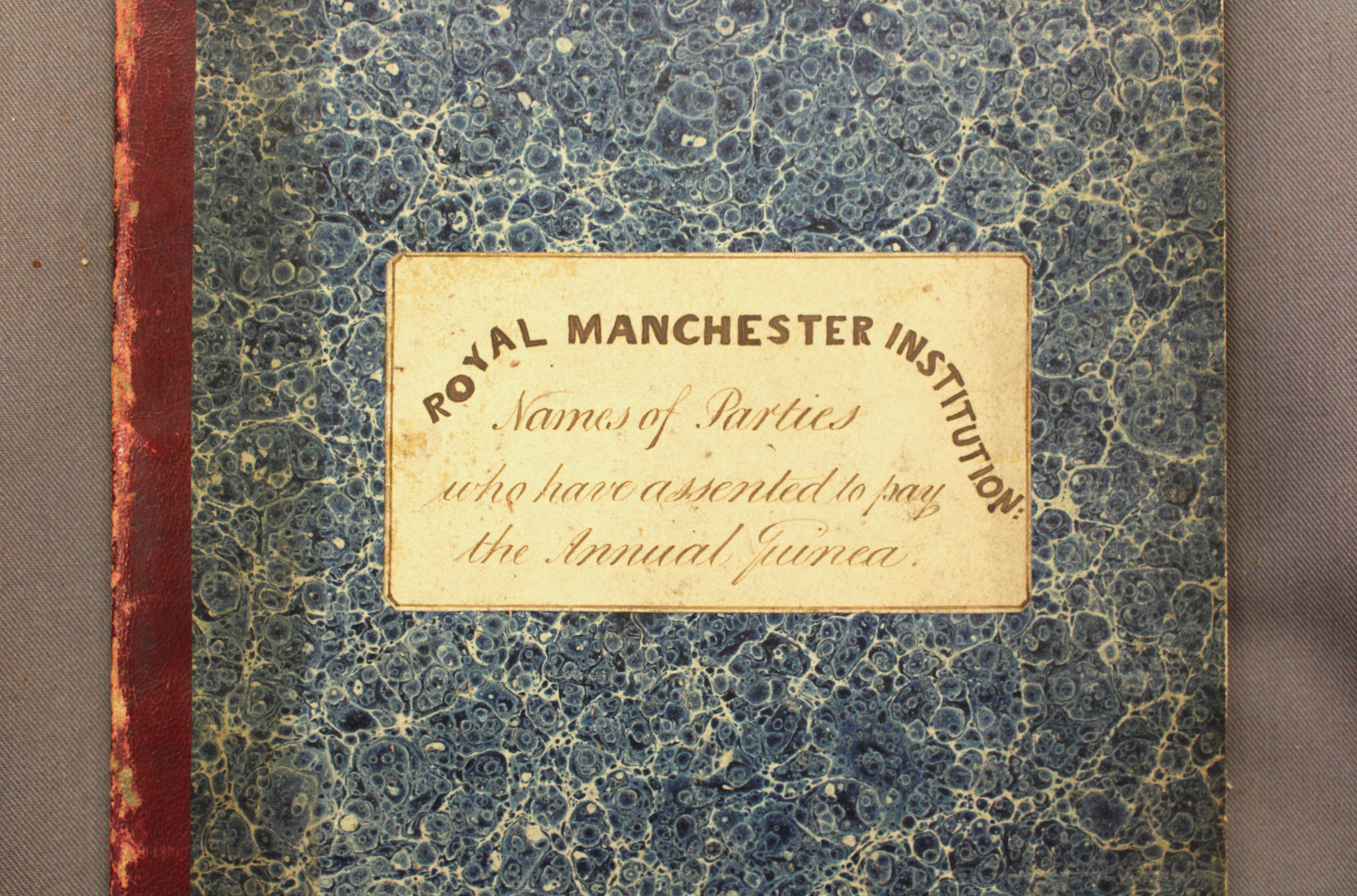 A cropped image showing the middle portion of the front cover of a large book. The cover is decorated with blue speckled marbled paper, and has a dark red leather binding along its spine. A white sticker on the middle of the front cover reads: ‘Royal Manchester Institution: Names of Parties who have Assented to Pay the Annual Guinea’. The book rests on grey canvas material – an archive book cushion.