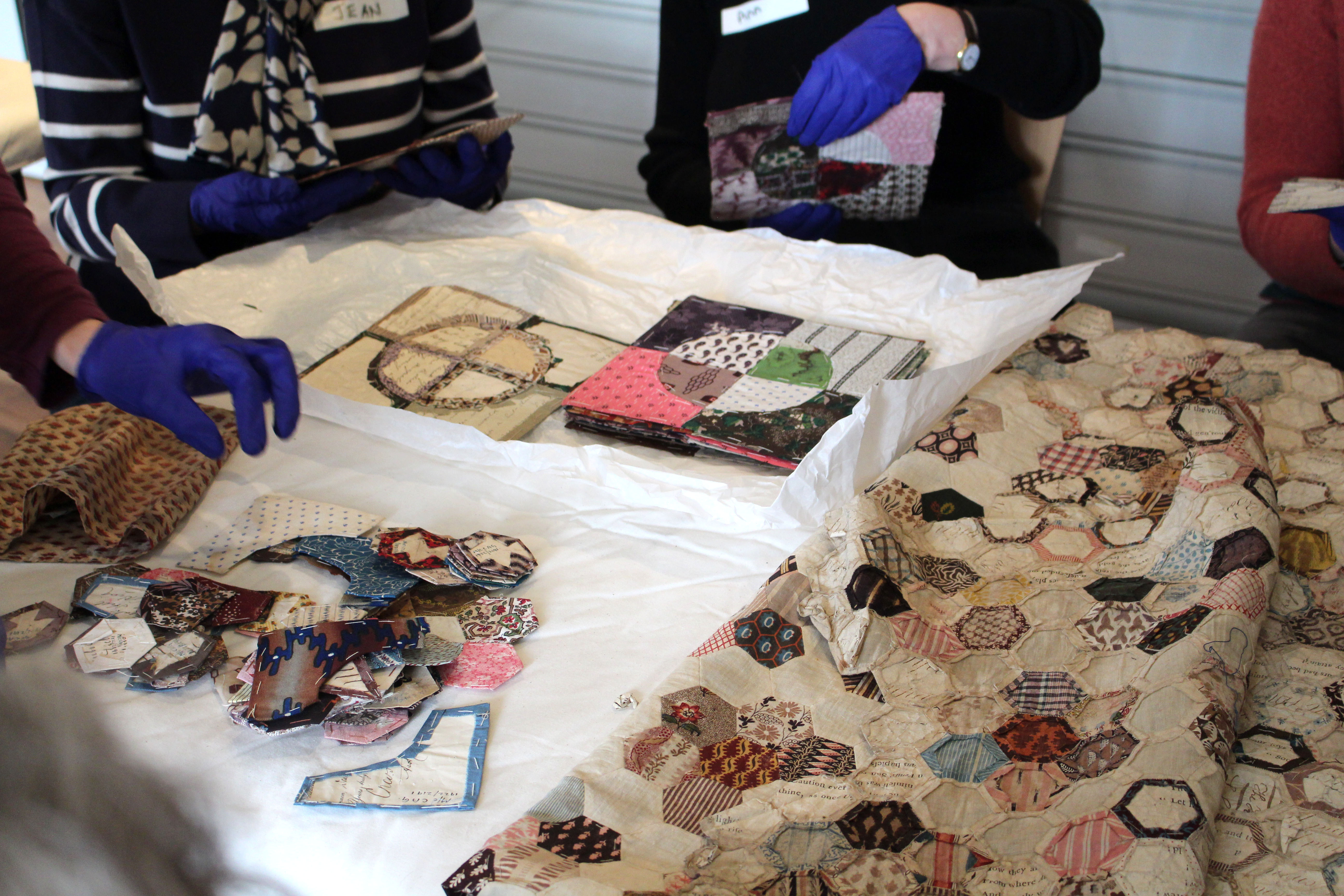 A table with a patchwork quilt and smaller patchwork swatches are in the foreground of the image. Four figures on the peripheries of the image, half cut off by the photograph’s frame, surround the table and handle some of the smaller samples with purple-gloved hands.