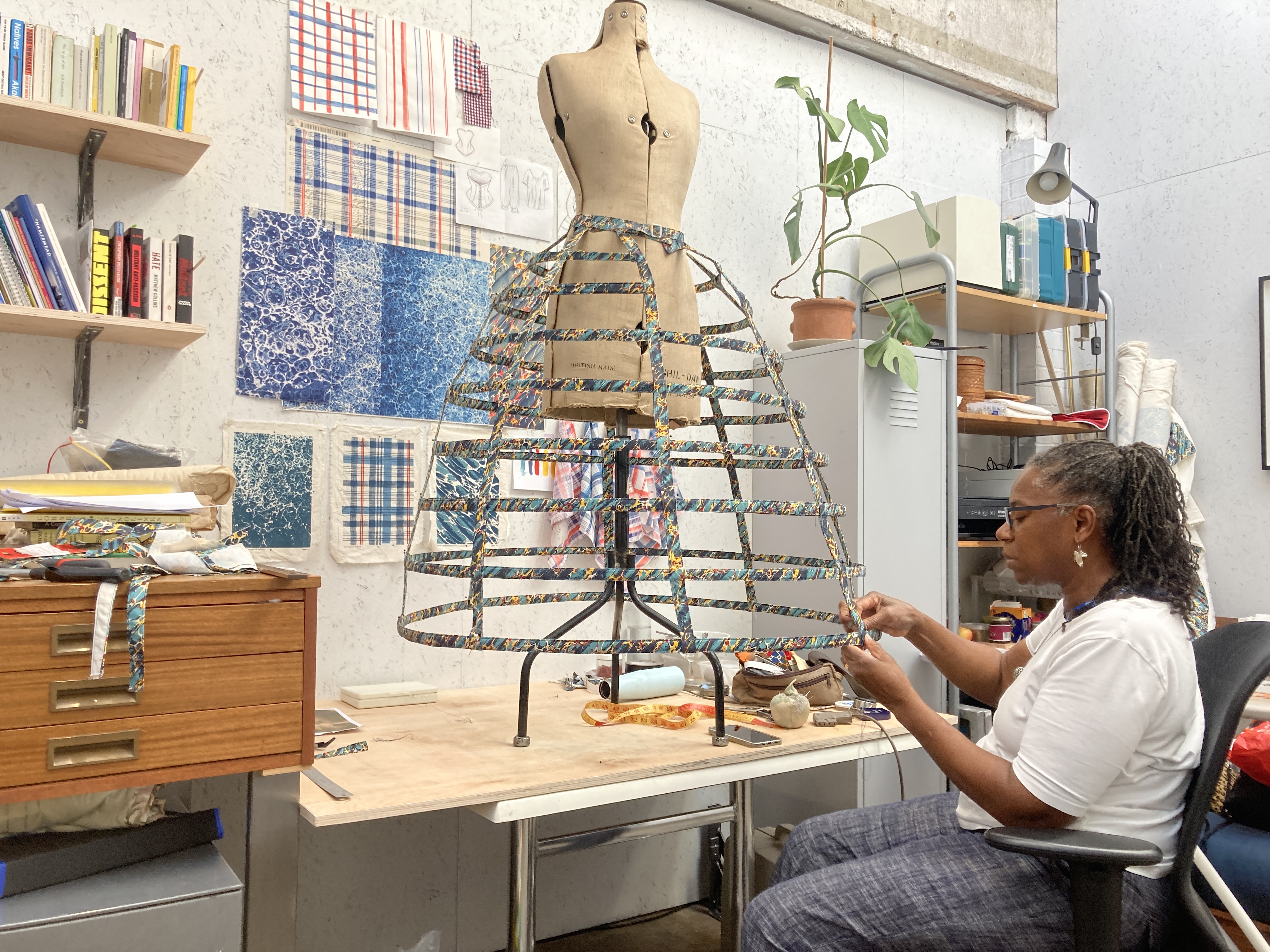 A studio space with shelves of books and designs on the wall. In the centre of the image is a table holding a dressmakers’ mannequin, wearing a crinoline. A seated woman to the right of the image stitched the hem of this crinoline.