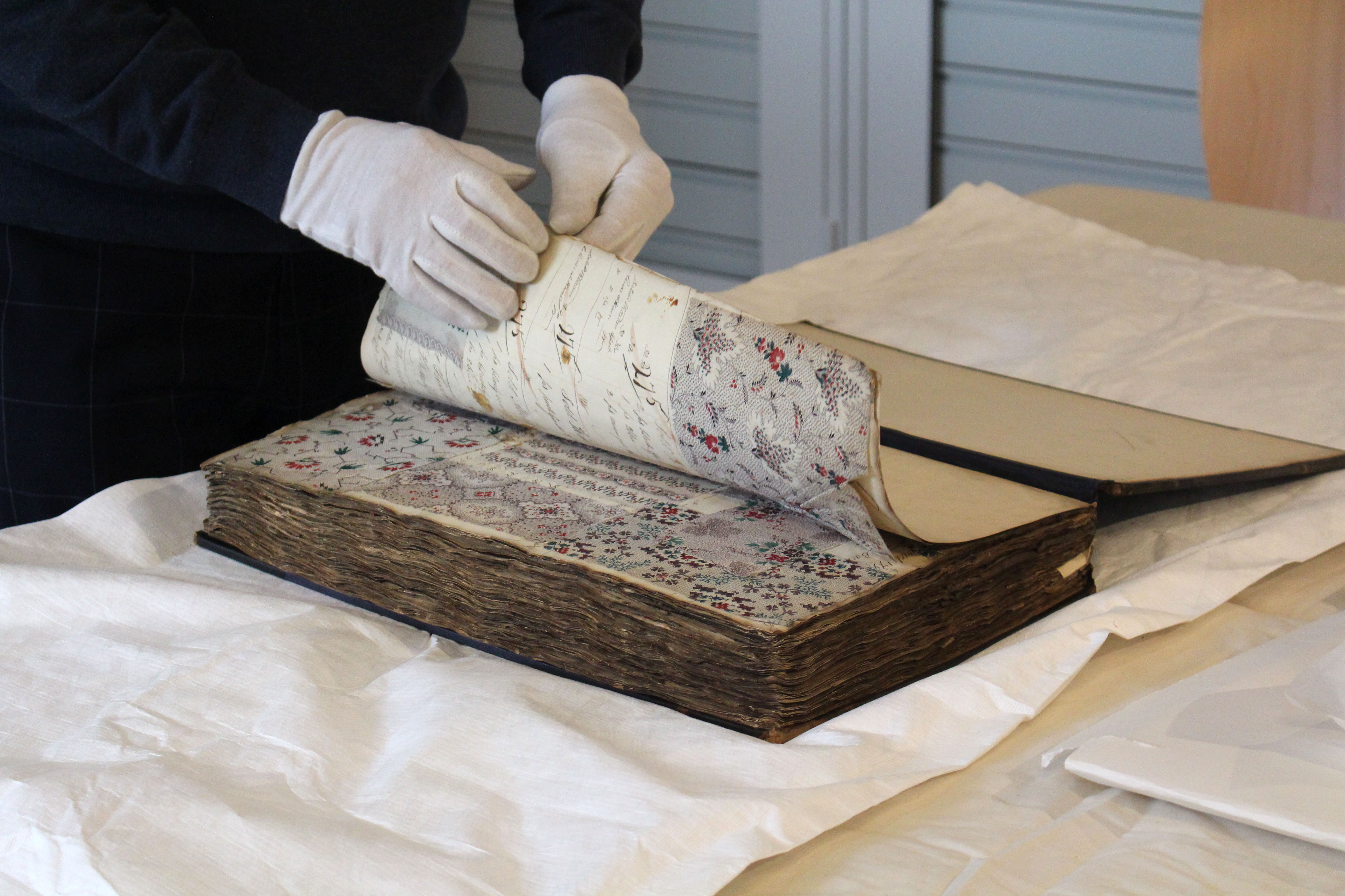 Two white gloved hands leaf through an antique ledger containing floral patterns.