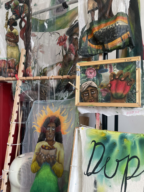 A studio space showcasing multiple mixed-media artworks. In the centre, a wooden frame holds a painting of a face and hand grasping a red plant pod. To the left, a translucent fabric artwork features a woman holding a bowl with a fiery golden aura surrounding her head. Behind and above, additional painted fabric works depict vibrant plants, flowers, and figures. The scene is layered, with an unfinished wooden ladder and fabric pieces adding texture and depth. A green and white painted cloth with “DUP” in bold black letters is partially visible in the foreground.