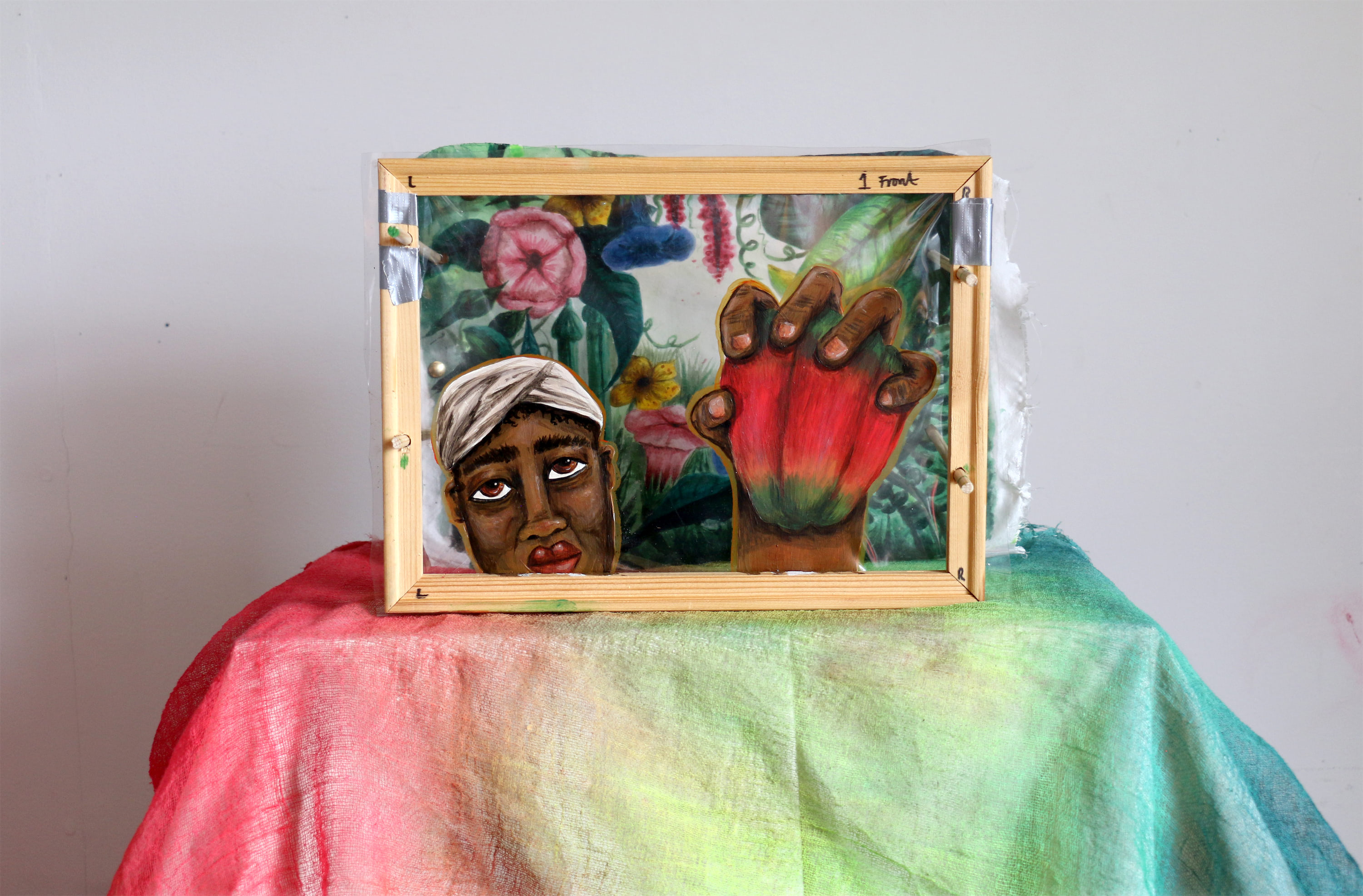 A wooden frame holding a painted artwork of a person’s face and hand. The hand holds a large, red and green plant resembling a pod. The background features intricate plants, including pink and blue flowers.