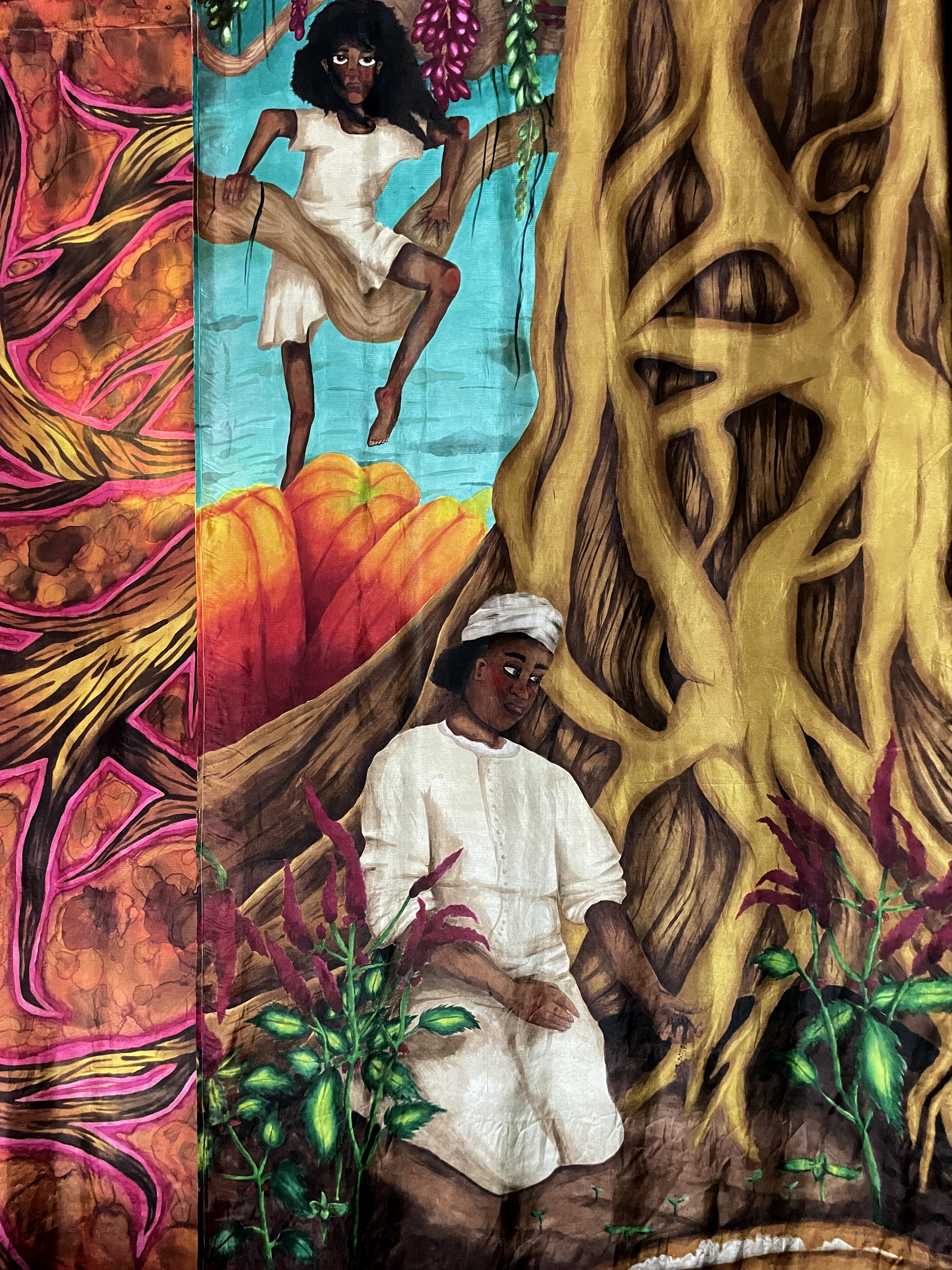 Close-up of a textile artwork showing two Black female figures interacting with an oversized tree. One is perched on a thick branch, looking outward with a firm expression. The other is kneeling at the base of the tree, dressed in white, with plants growing around her. The background includes pumpkin-like forms and vivid turquoise sky, with roots of the tree spreading into the earth.