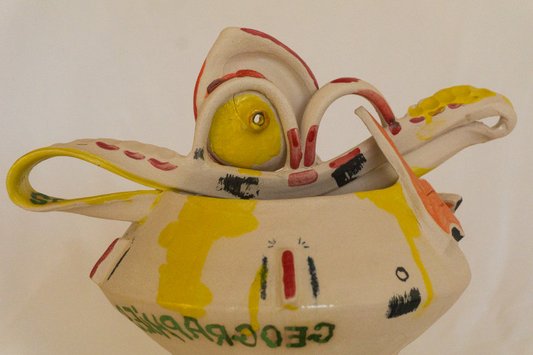 A painted ceramic piece with curved extensions and intricate shapes, featuring a yellow painted “eye” and green text reading “GEOGRAPHY.” The sculpture is covered with red, yellow, and black abstract markings.