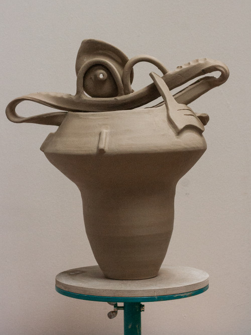 A raw clay ceramic piece on a pottery wheel. The sculpture has intricate, curving elements at the top resembling abstract shapes, still in its unpainted stage.