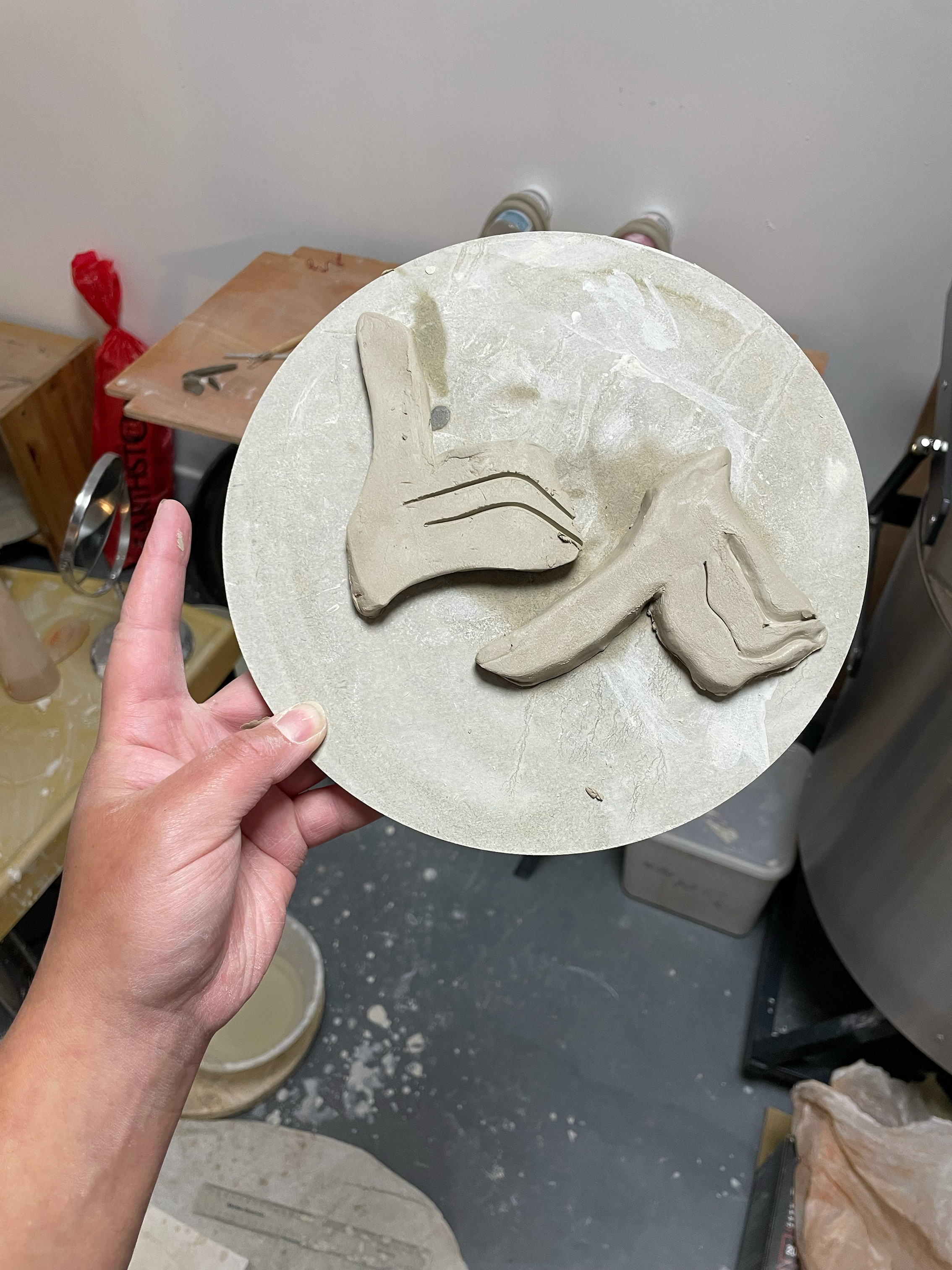 A clay piece featuring abstract shapes on a round ceramic plate. The hand holding the plate shows the scale of the sculpture, with tools and equipment visible in the workshop background.