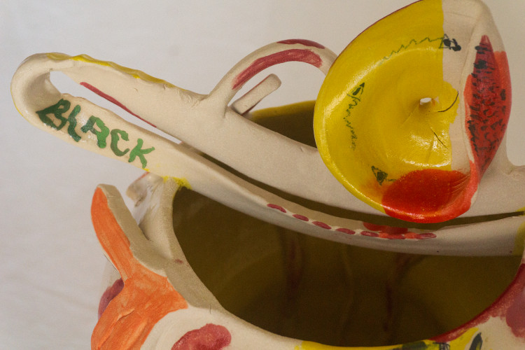 A close-up view of a ceramic sculpture featuring bold yellow, red, and orange painted patterns. A prominent handle-like structure is painted with the word “BLACK” in green.