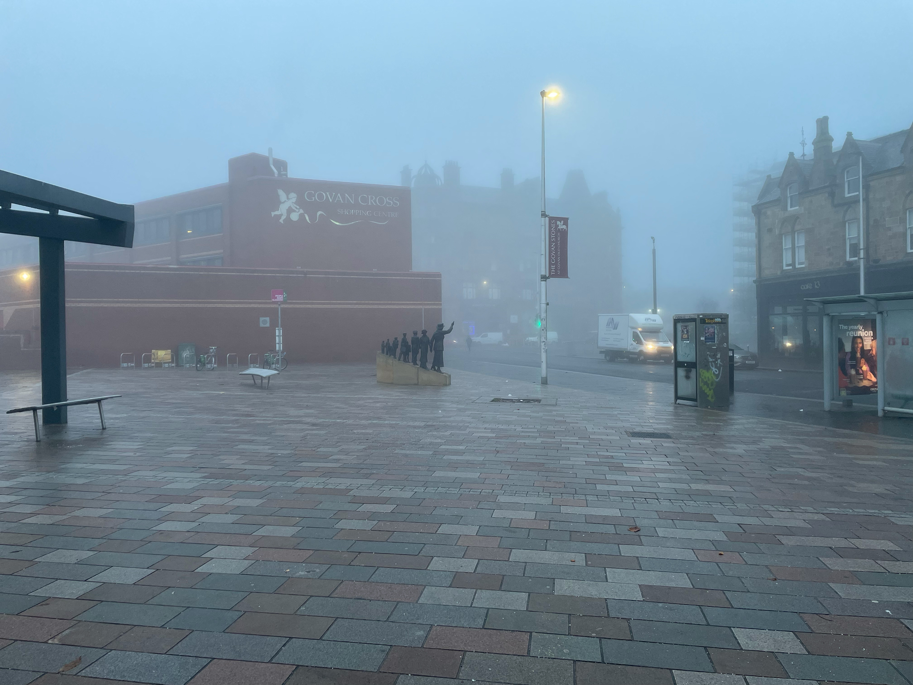 A misty view of a city square near “Govan Cross Shopping Centre.” The foreground has wet brick pavement, and the area appears quiet, with silhouettes of statues and a few vehicles in the distance.
