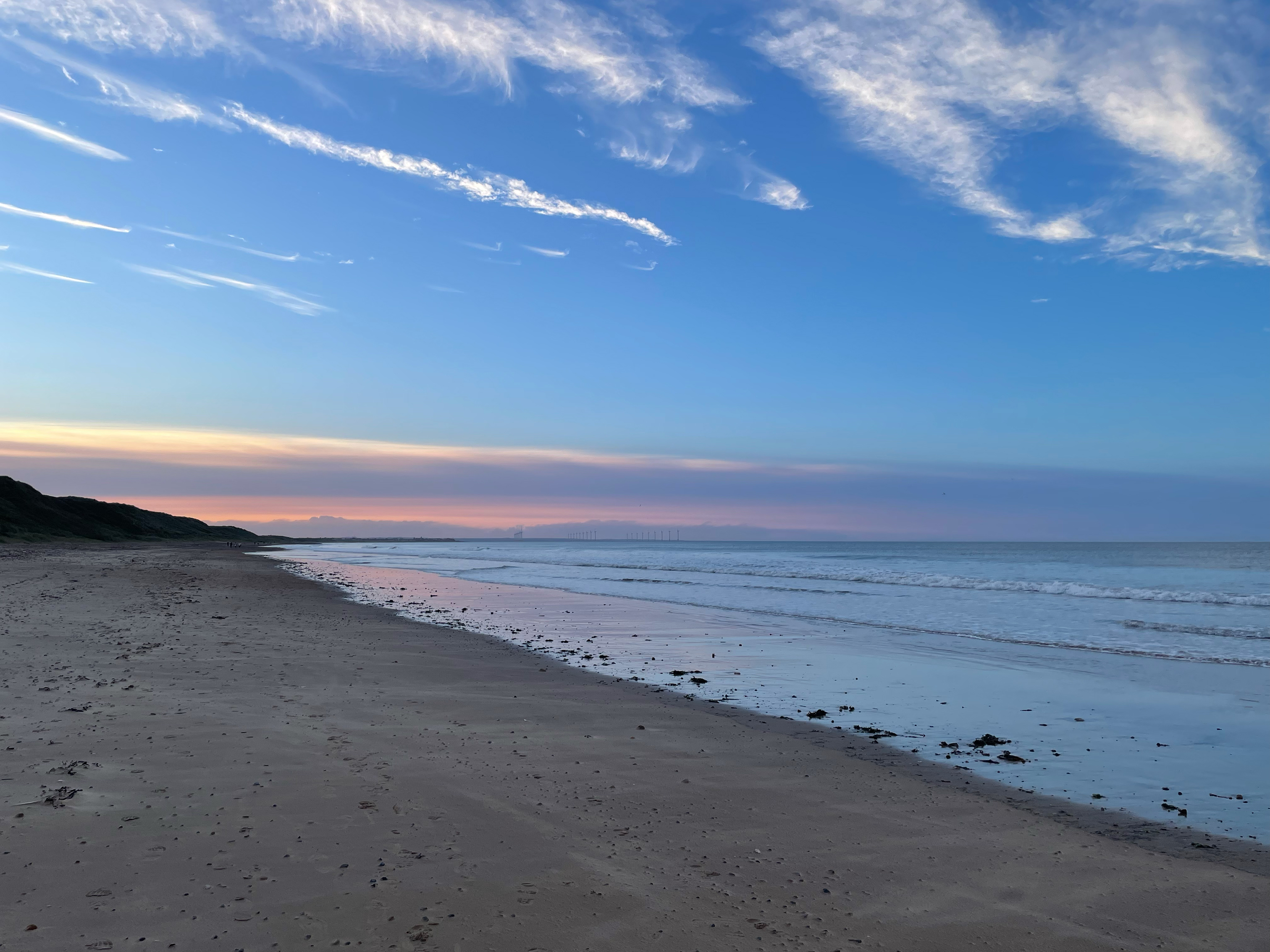 A calm sandy beach with the ocean to the right and a vibrant sky filled with blue, pink, and orange hues. Waves gently lap the shore, and the coastline stretches into the distance with a silhouette of dunes or hills.